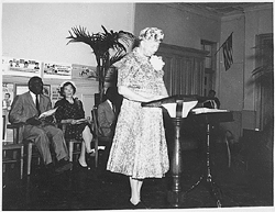 Eleanor Roosevelt speaking to an audience at the Manhattan School for Boys. Jackie Robinson, along with other guests, are seated directly behind Roosevelt as she speaks.